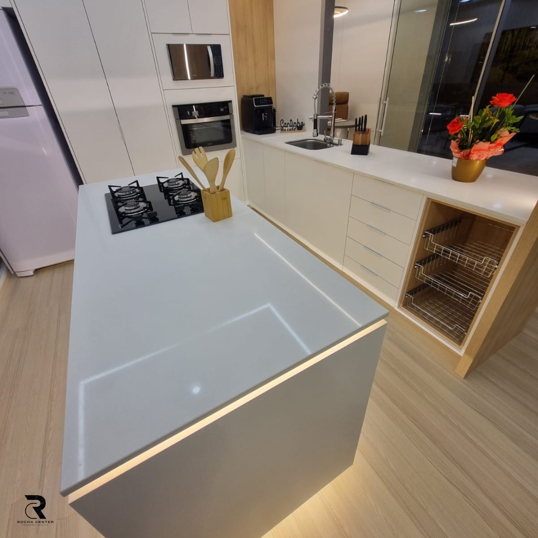 Imagem de uma cozinha moderna com iluminação, mostrando uma ilha de cozinha branca com gás, bancada de trabalho e armários brancos e madeira